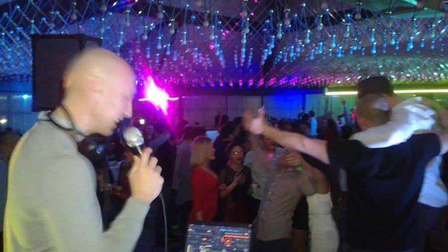tomislav 3 DJ Tomislav (Tom Black) drži mikrofon dok vodi program na vjenčanju, s osmijehom na licu ispred DJ pulta.